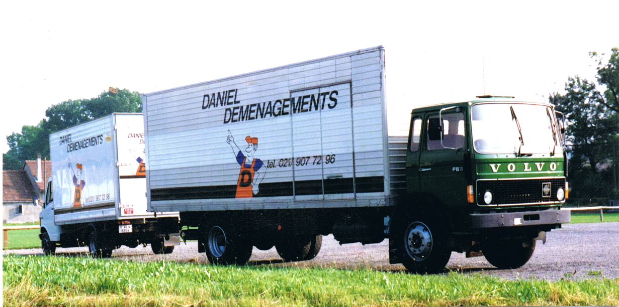 Deux camions de déménagement garés devant une maison, des déménageurs chargeant des meubles. Journée ensoleillée, ciel bleu.
