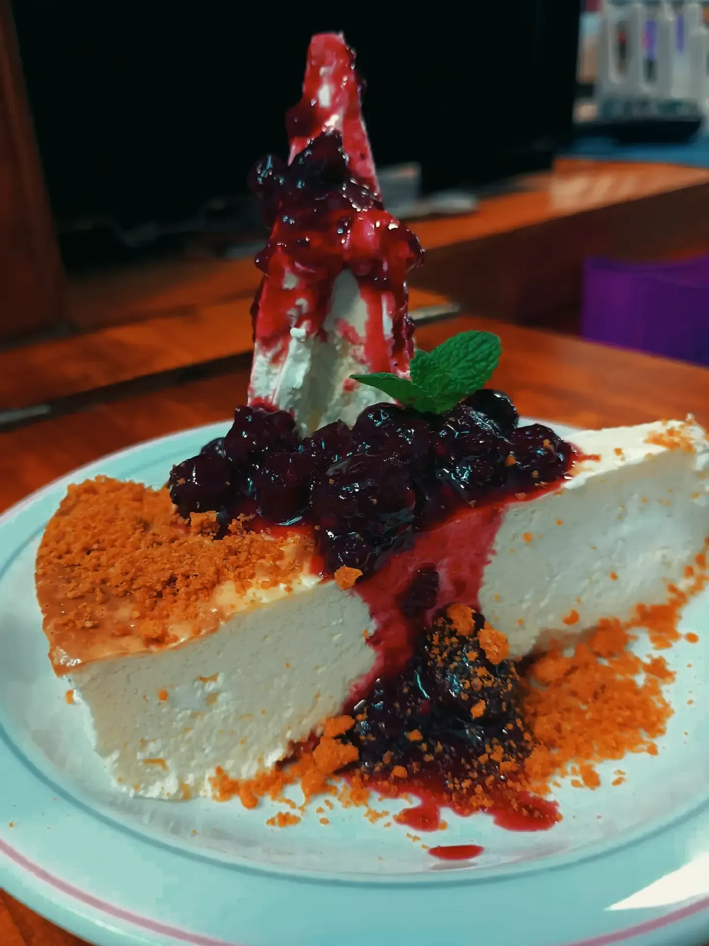 Rebanada de tarta de queso con cobertura de frutos rojos, crema batida y crumble de galletas en un plato blanco.