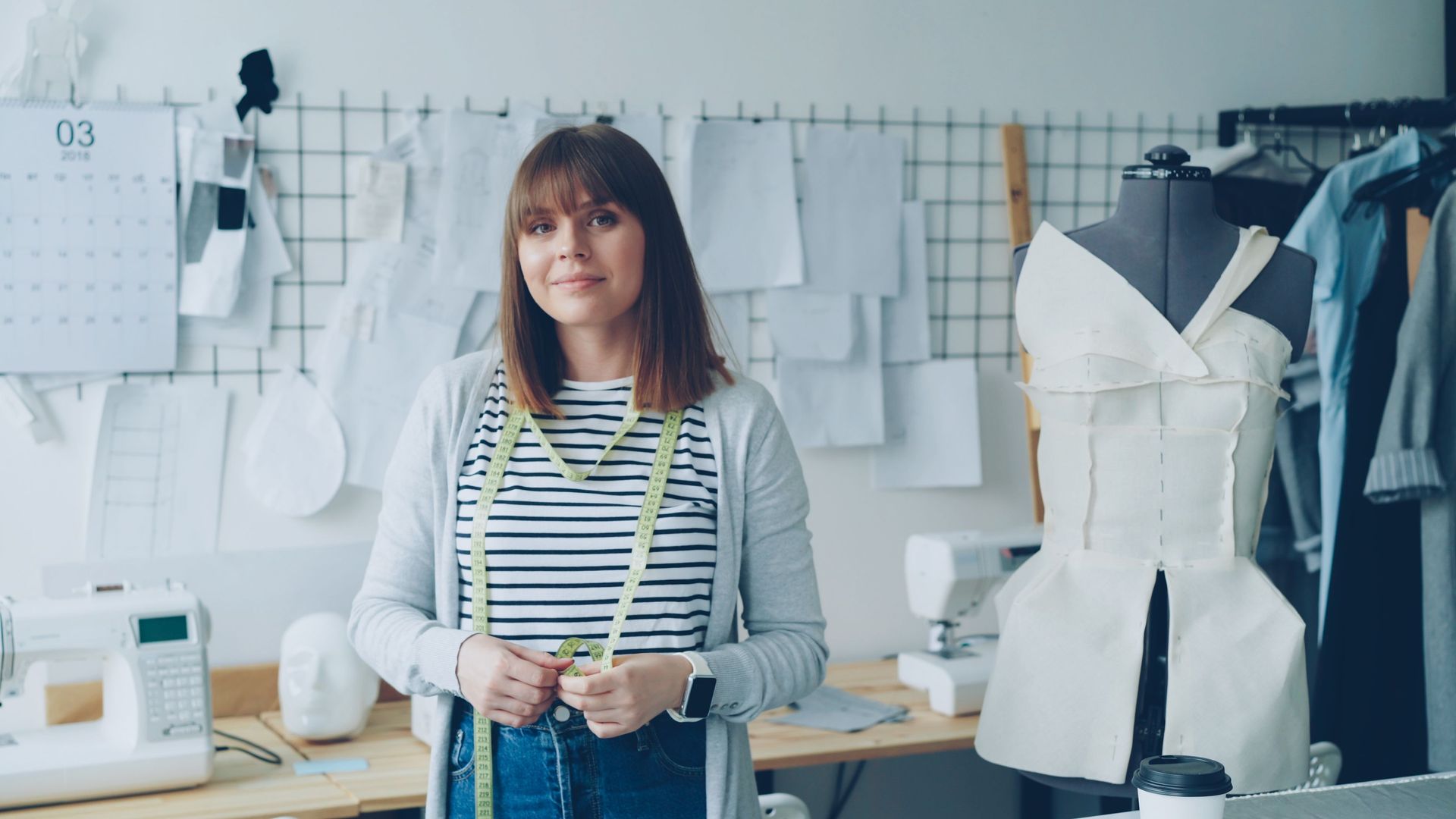 Una persona de pie en un estudio de diseño de moda junto a bocetos de vestidos colgados y un vestido blanco sobre un maniquí.
