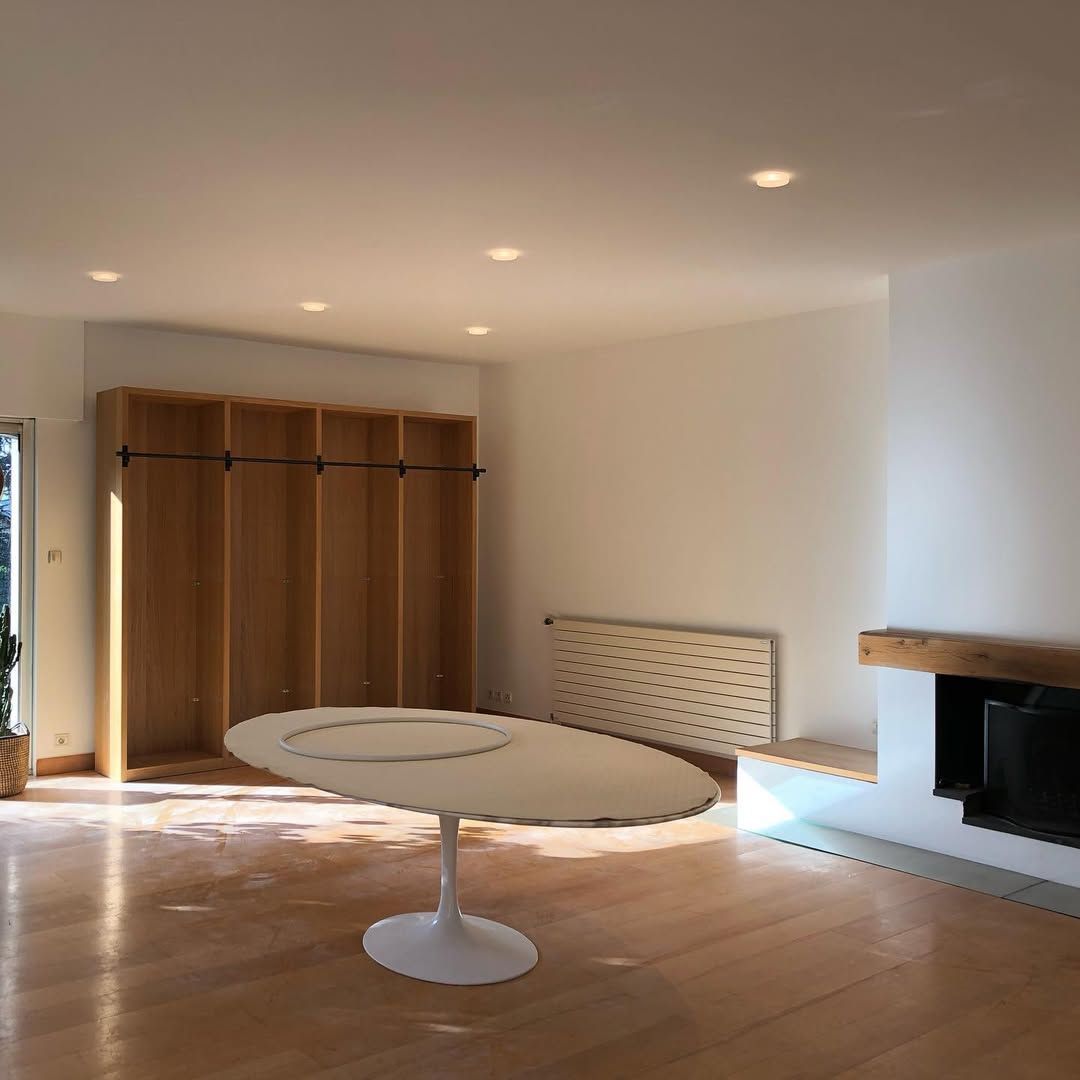 Salon vide et ensoleillé avec parquet, étagères intégrées, cheminée et table à manger ovale blanche.