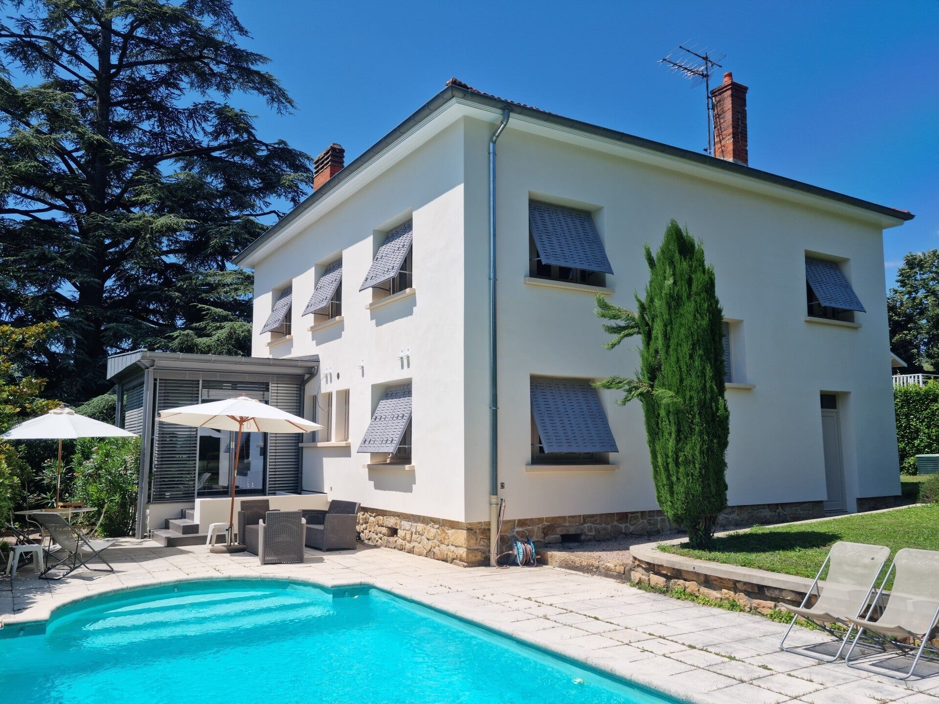 Maison blanche avec piscine, volets bleus et mobilier de jardin.