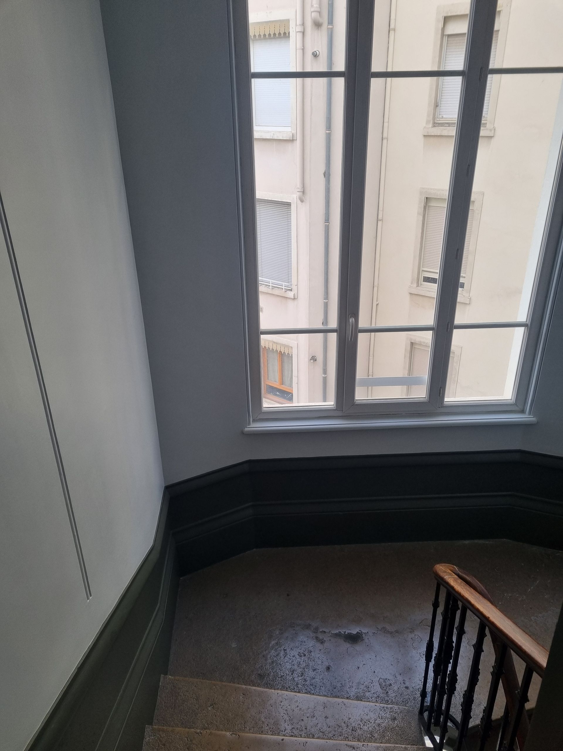 Cage d'escalier avec murs gris, rampe en bois et fenêtre donnant sur les bâtiments.