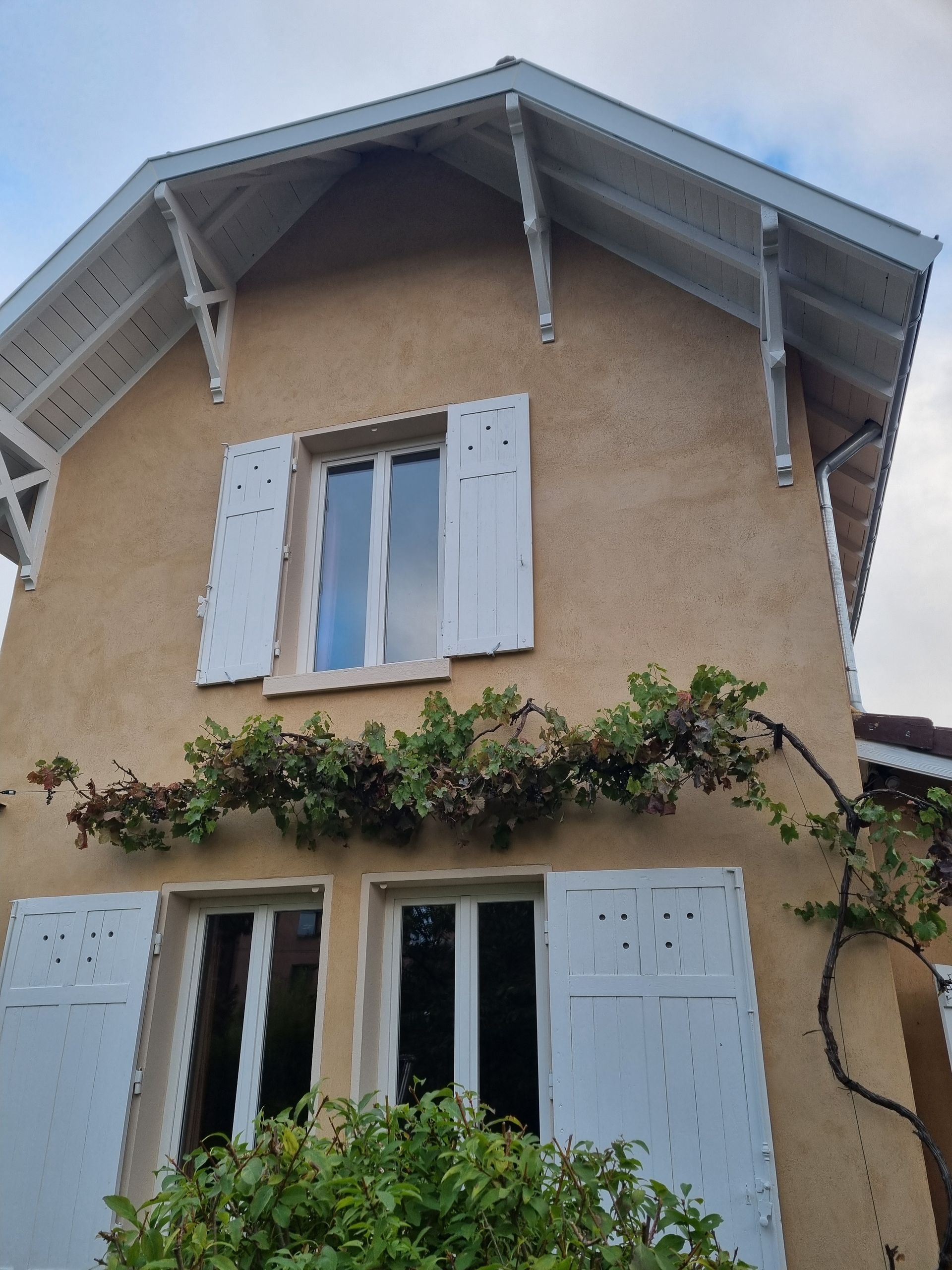 Maison beige aux volets et boiseries blanches. Une vigne grimpe le long du mur.