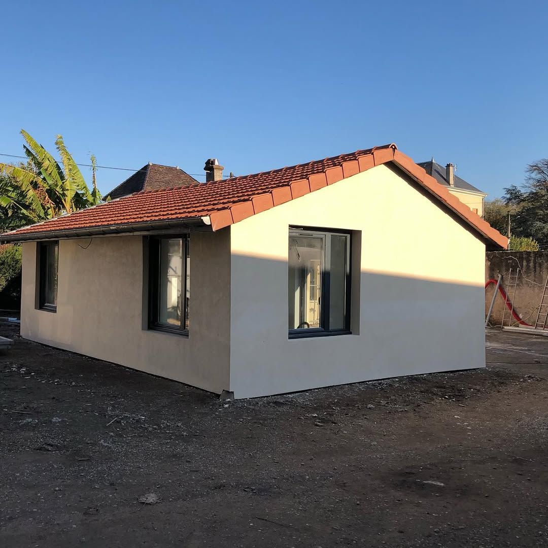 Petit bâtiment de couleur crème avec un toit en tuiles de terre cuite, deux fenêtres, posé sur du gravier, sous un ciel bleu.