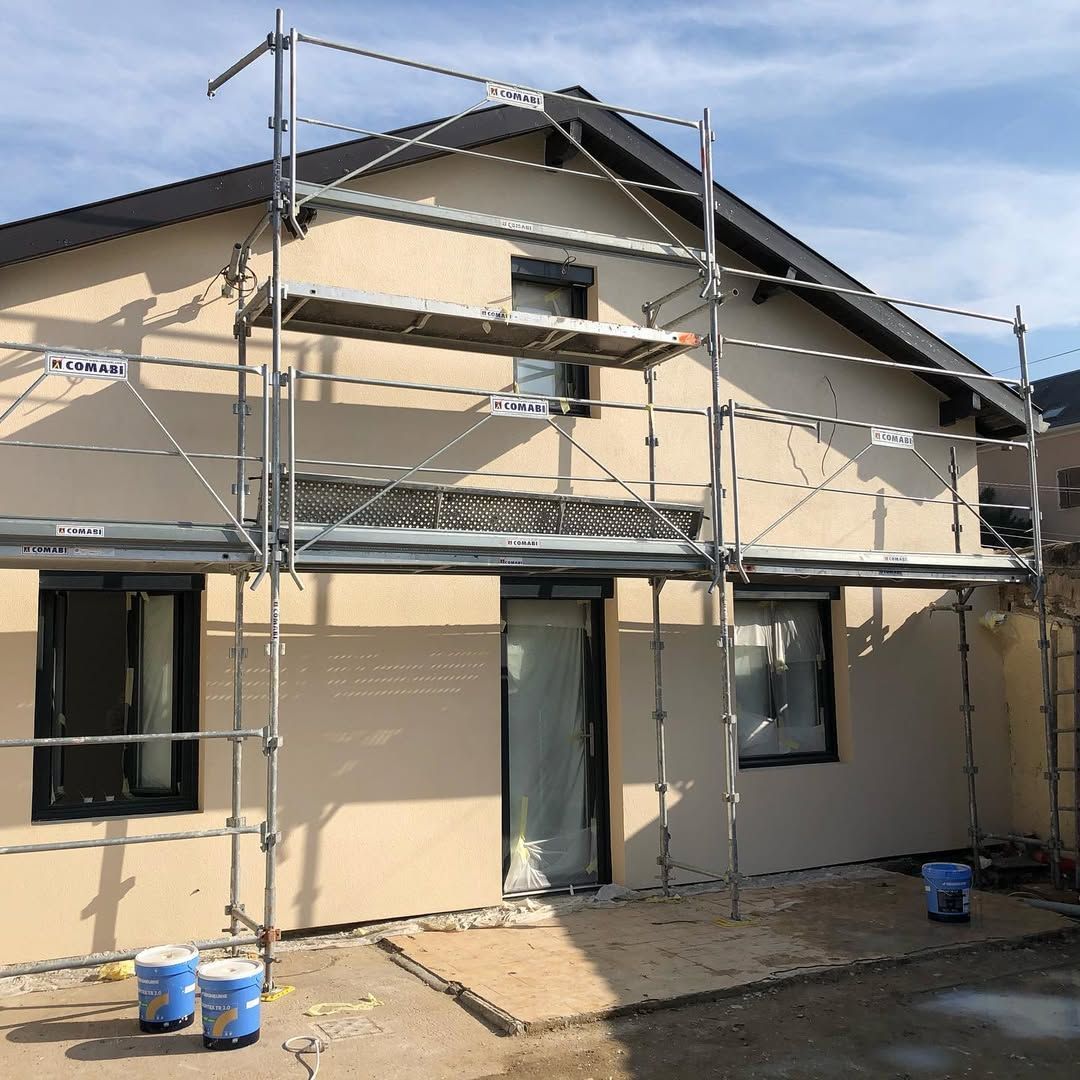 Échafaudage sur une maison en cours de rénovation, avec revêtement beige, garniture noire et seaux de peinture bleus.