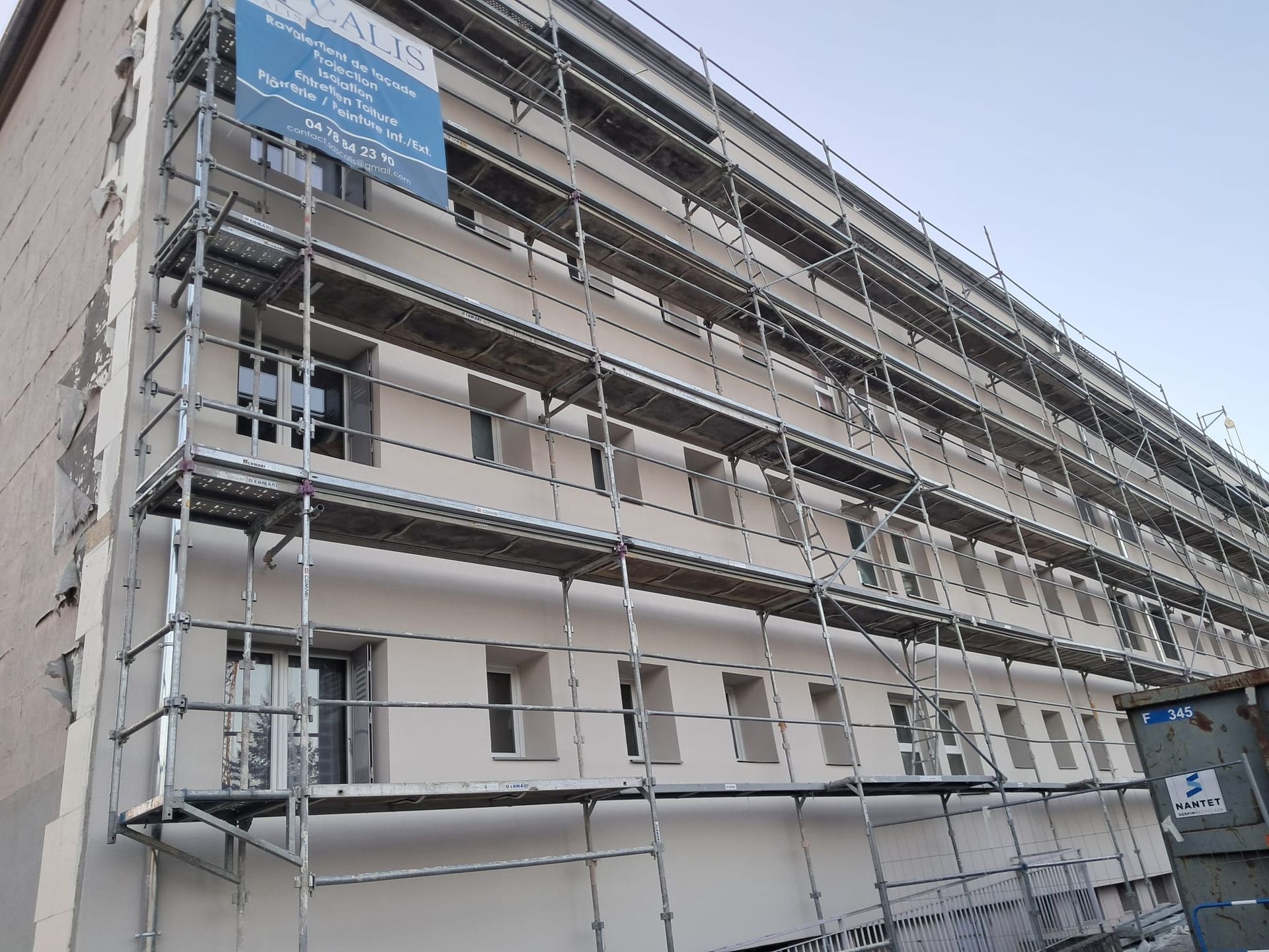 Échafaudages sur la façade du bâtiment en cours de rénovation, avec des fenêtres visibles et un panneau bleu.