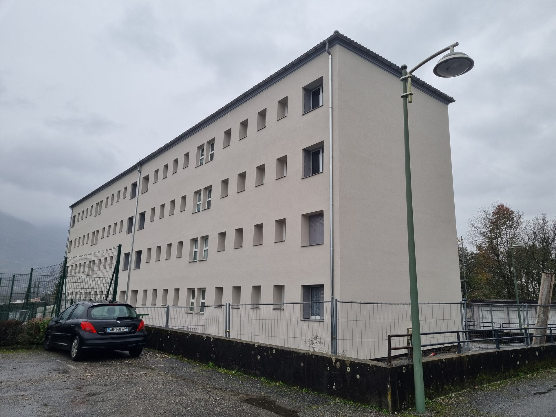 Immeuble beige de trois étages avec des rangées de fenêtres, voiture garée devant, ciel couvert.