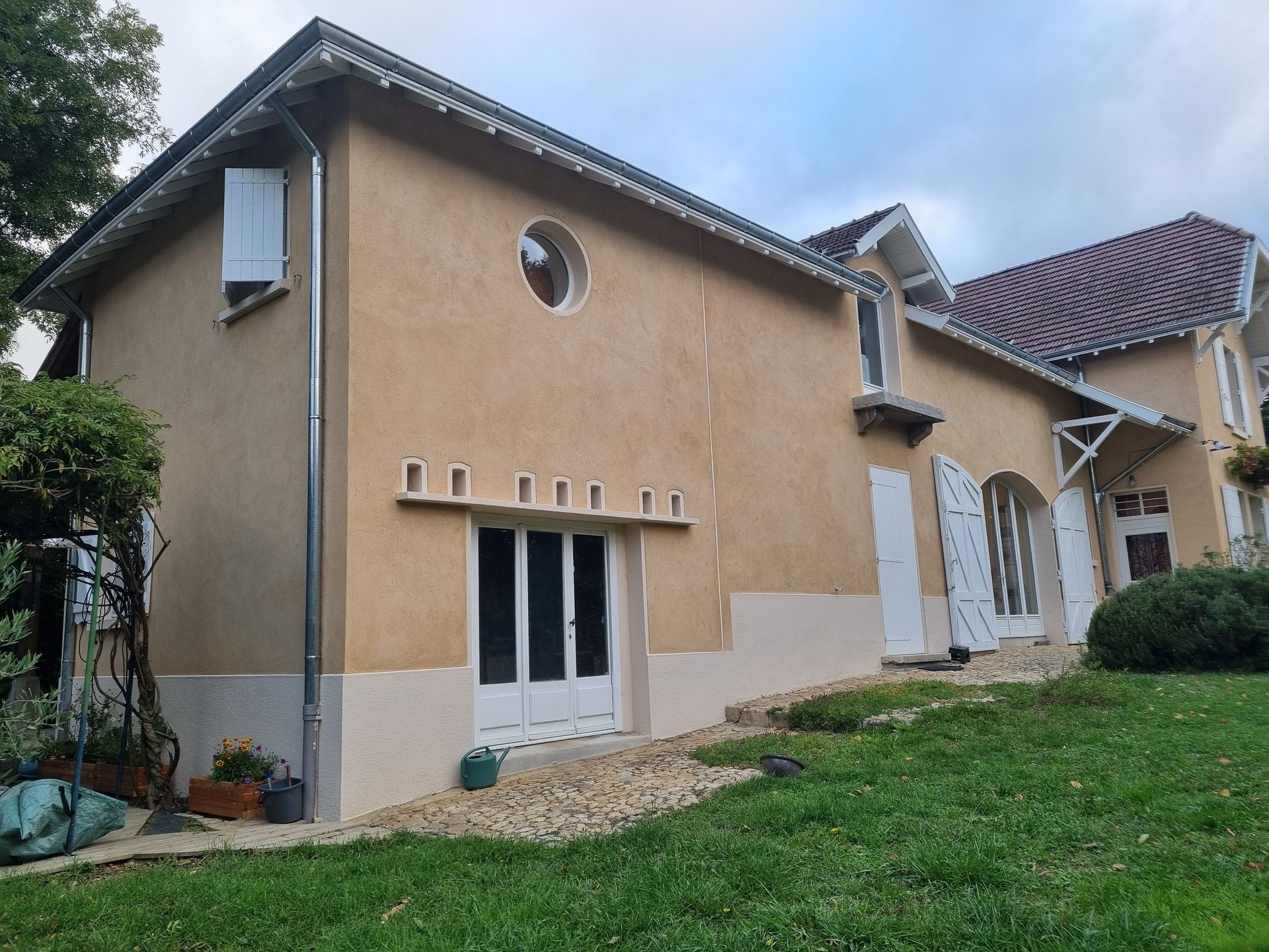 Maison en stuc beige avec des boiseries blanches, des fenêtres cintrées et un toit de tuiles rouges sous un ciel nuageux.