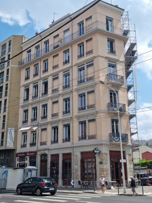 Immeuble beige de sept étages avec balcon d'angle, café et échafaudages. Une voiture et des piétons se trouvent dans la rue.