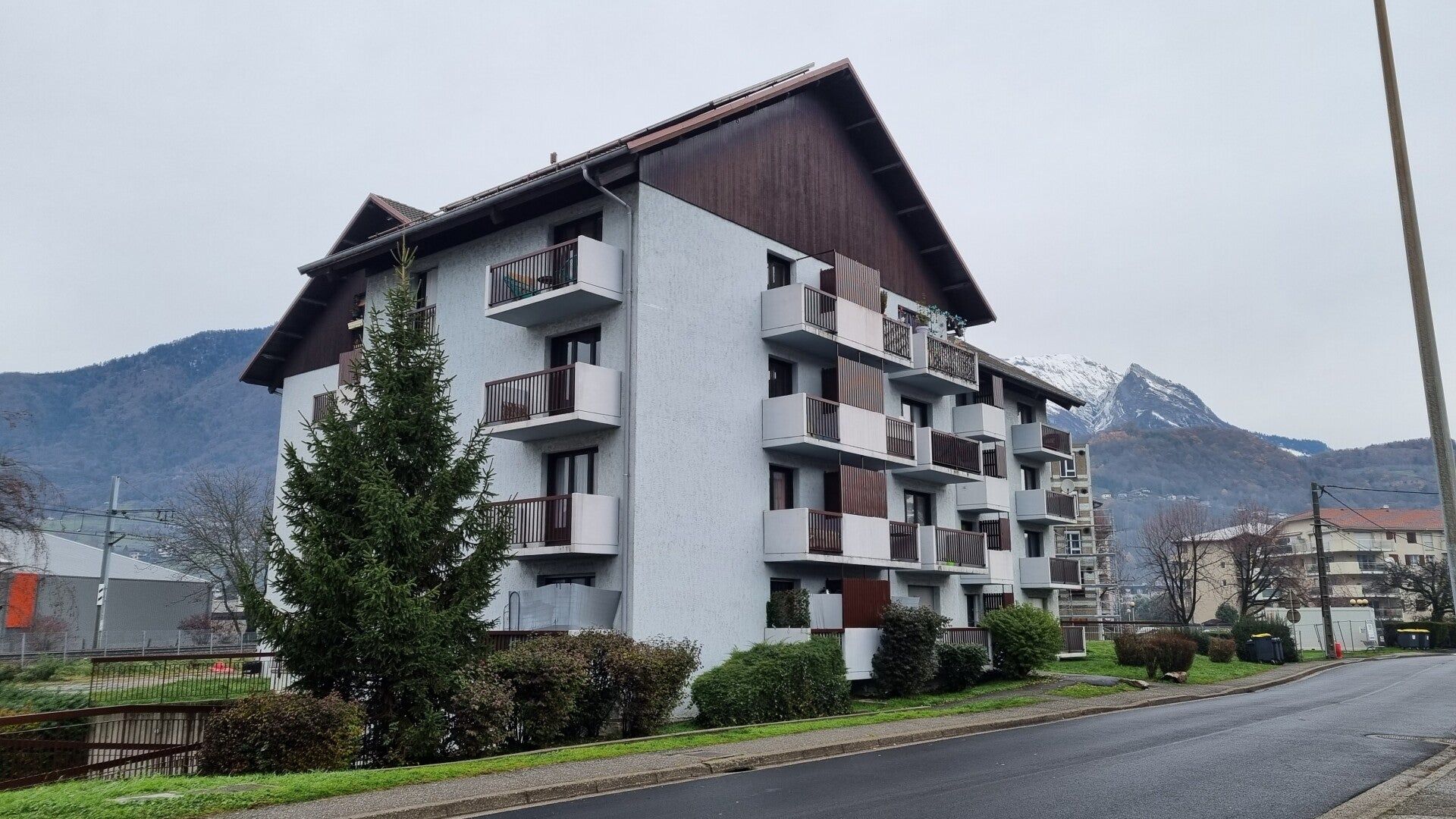 Immeuble d'appartements à plusieurs étages avec balcons, toiture brune et arrière-plan montagneux.