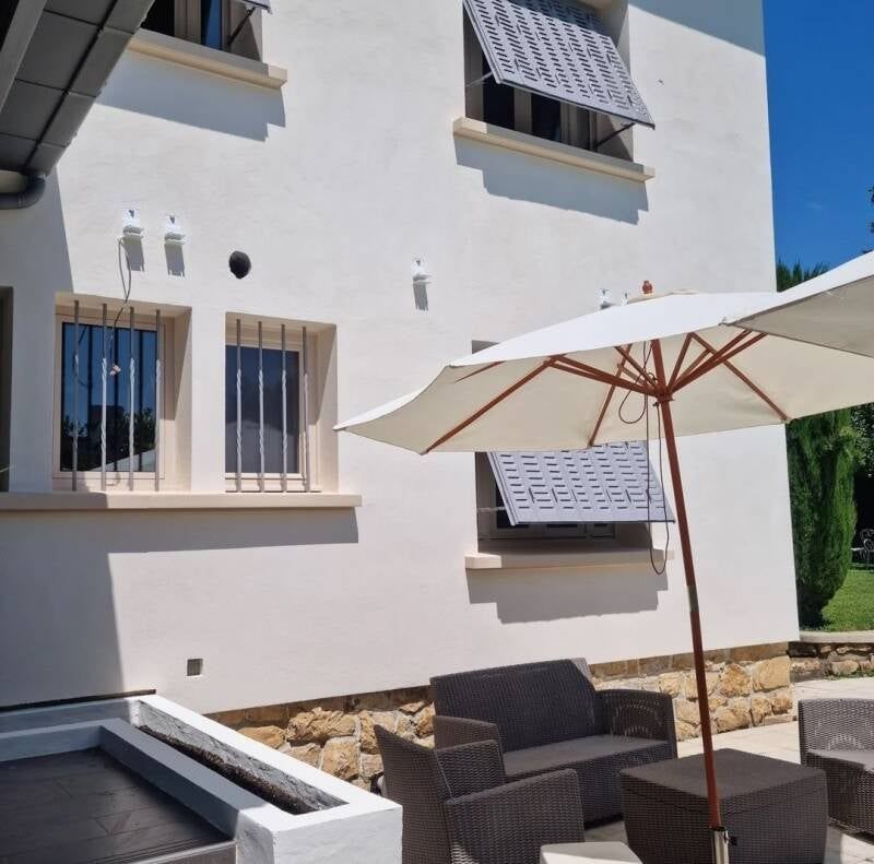 Extérieur d'une maison blanche avec mobilier de patio, parasol et fenêtres partiellement ombragées.
