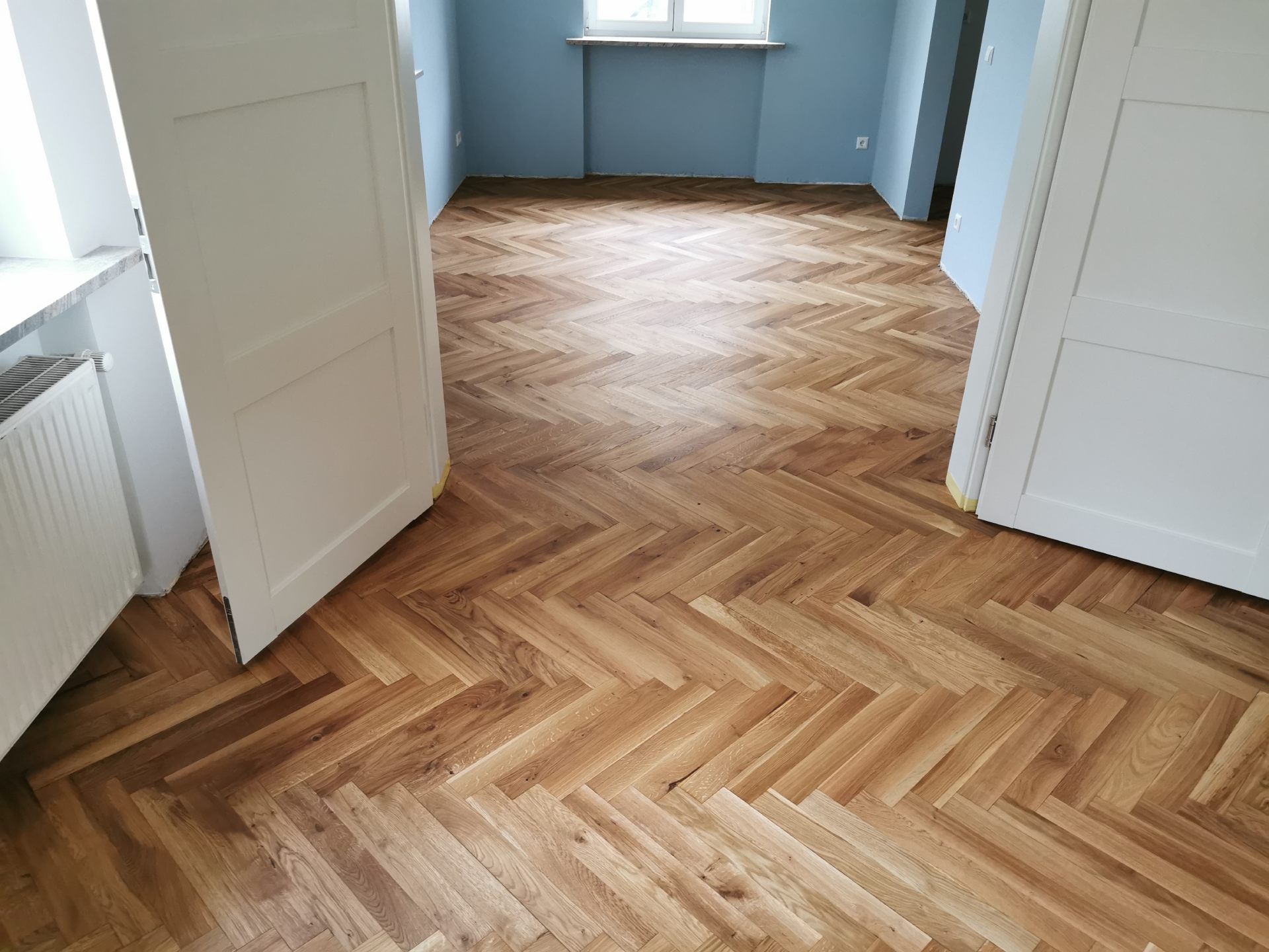 Ein Zimmer mit hellbraunem Fischgrätparkett, weißen Doppeltüren an beiden Seiten und hellblauen Wänden.