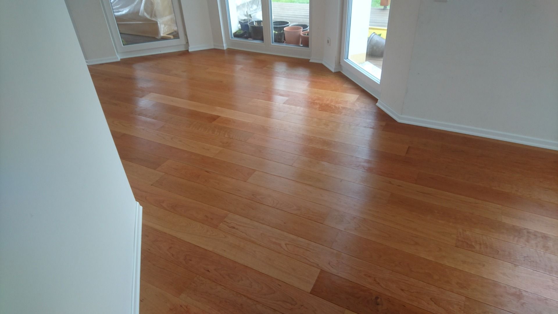 Ein heller, leerer Raum mit poliertem, mittelbraunem Hartholzboden und weißen Wänden, im Hintergrund Glastüren.