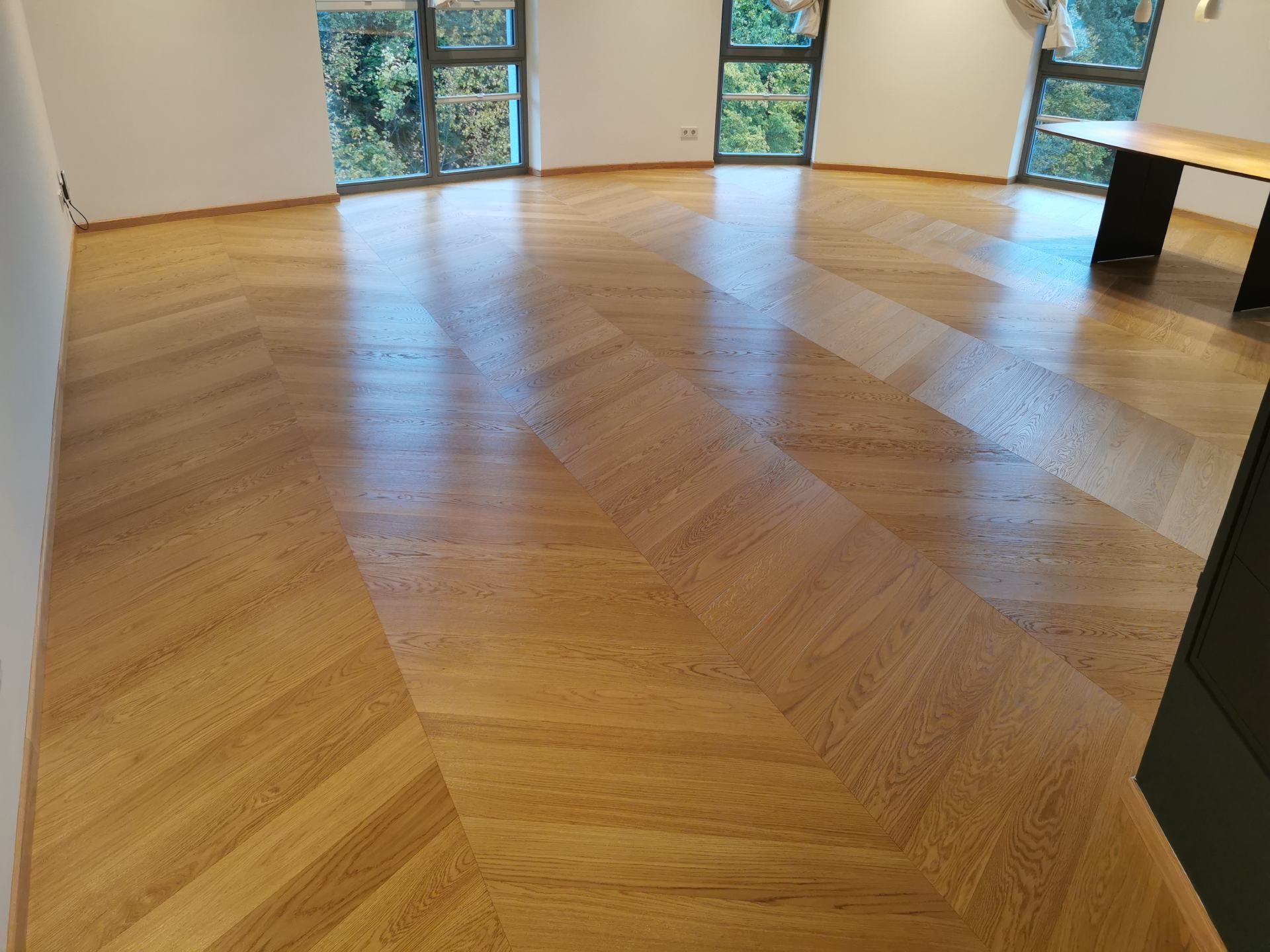 Ein Zimmer mit hellem, im Fischgrätmuster verlegtem Holzboden, weißen Wänden und großen Fenstern.