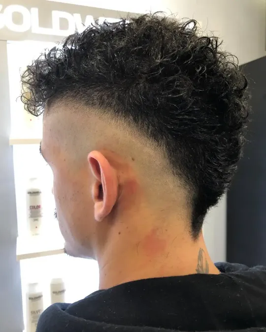 Vista lateral de un corte de pelo rizado con degradado alto, lados rapados y un tatuaje en el cuello en una barbería.