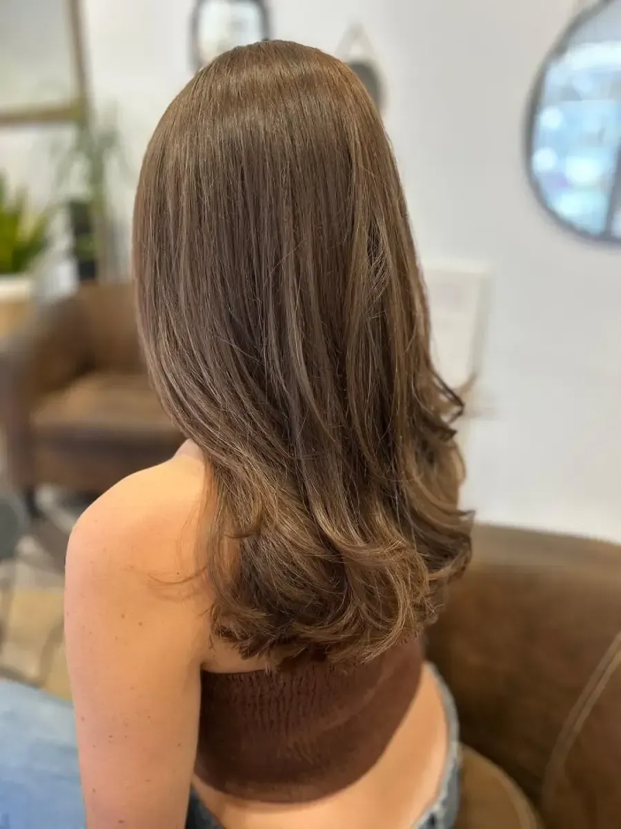 Vista trasera de una persona con cabello castaño largo, que lleva una blusa sin mangas en un salón de belleza.