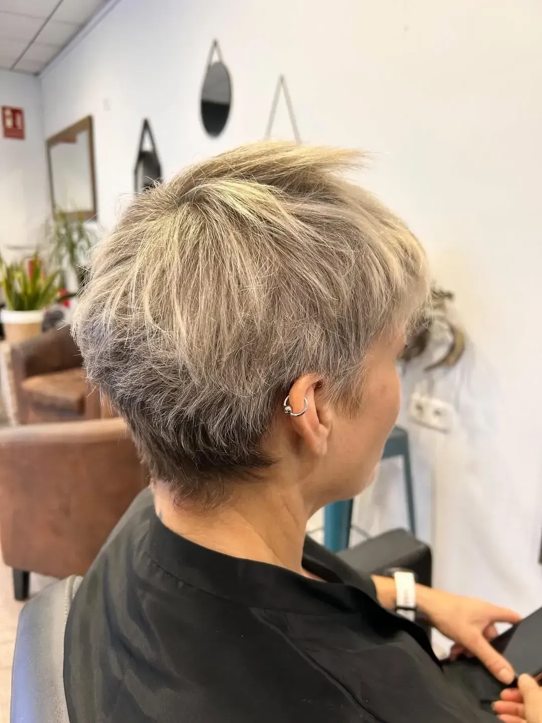 Corte de pelo corto, gris plateado y capeado, en una peluquería, vista lateral, en una persona sentada con una capa negra.