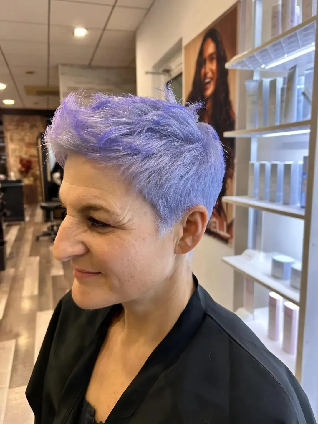 Persona con cabello corto color lavanda en una peluquería, mostrada de perfil con una leve sonrisa.