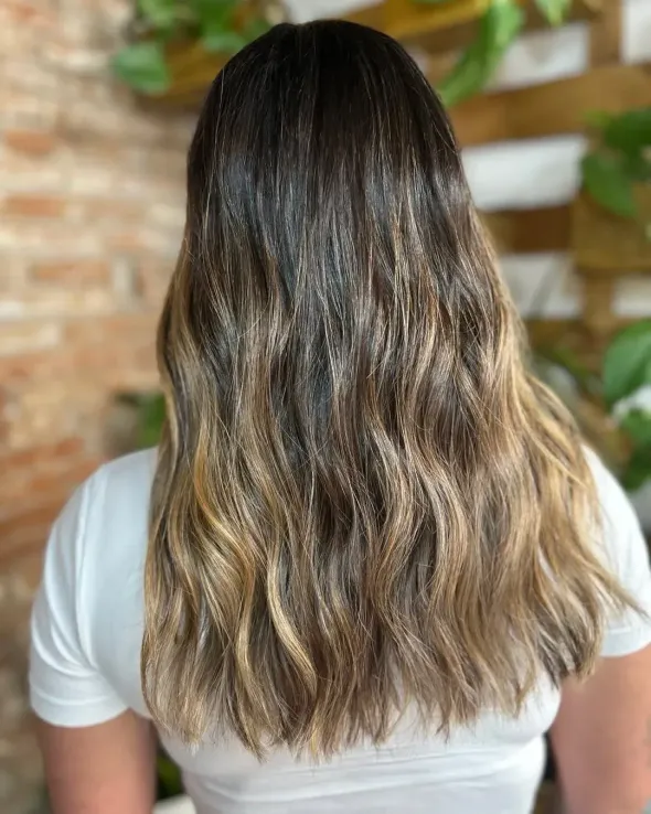 Vista posterior de una larga melena ondulada de color castaño oscuro con puntas rubias más claras sobre un fondo de ladrillos
