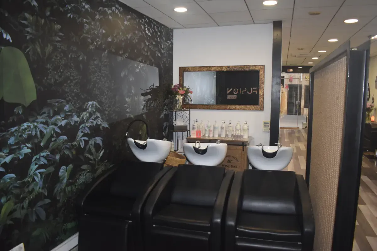 Tres lavabos negros para champú en un salón de belleza con un mural de hojas en la pared y un espejo enmarcado.