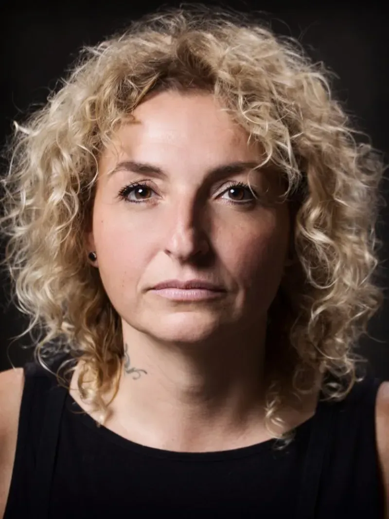 Retrato de una persona con cabello rubio rizado sobre un fondo oscuro, vestida con una blusa negra, mirando a la cámara.