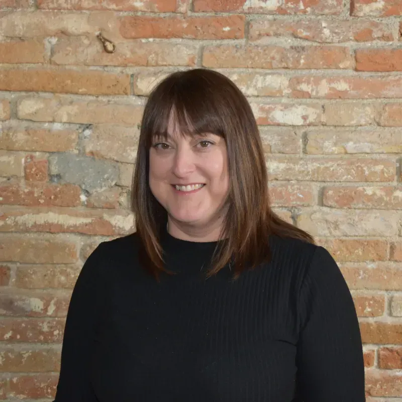 Persona sonriente con cabello castaño y blusa negra de pie contra una pared de ladrillos.