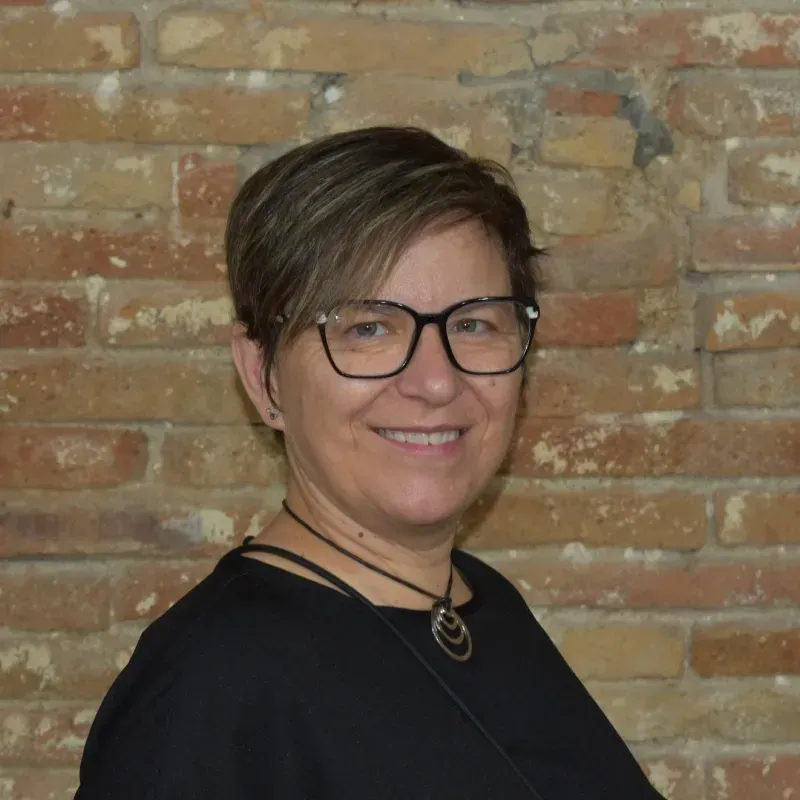 Persona con cabello corto y gafas sonriendo frente a una pared de ladrillos.