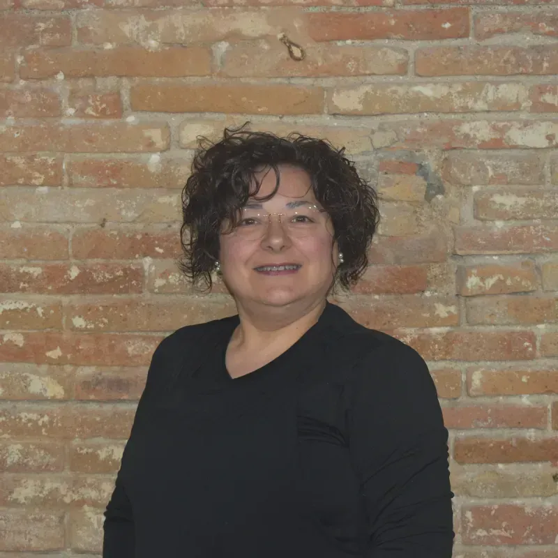 Persona con cabello oscuro y rizado, vestida con una blusa negra, de pie contra una pared de ladrillos.