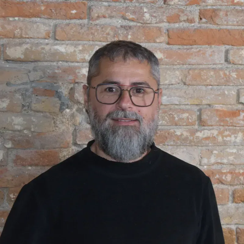 Retrato de un hombre con gafas y barba gris, vestido con una camisa negra, de pie contra una pared de ladrillos.