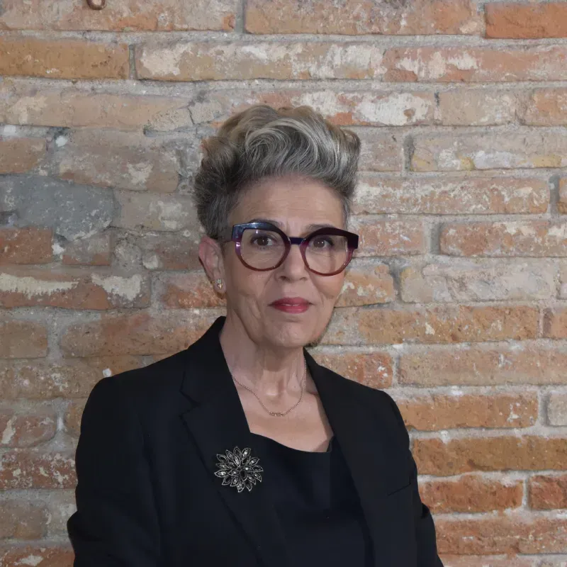 Mujer con cabello gris y gafas, vestida con un blazer negro, apoyada contra una pared de ladrillos.
