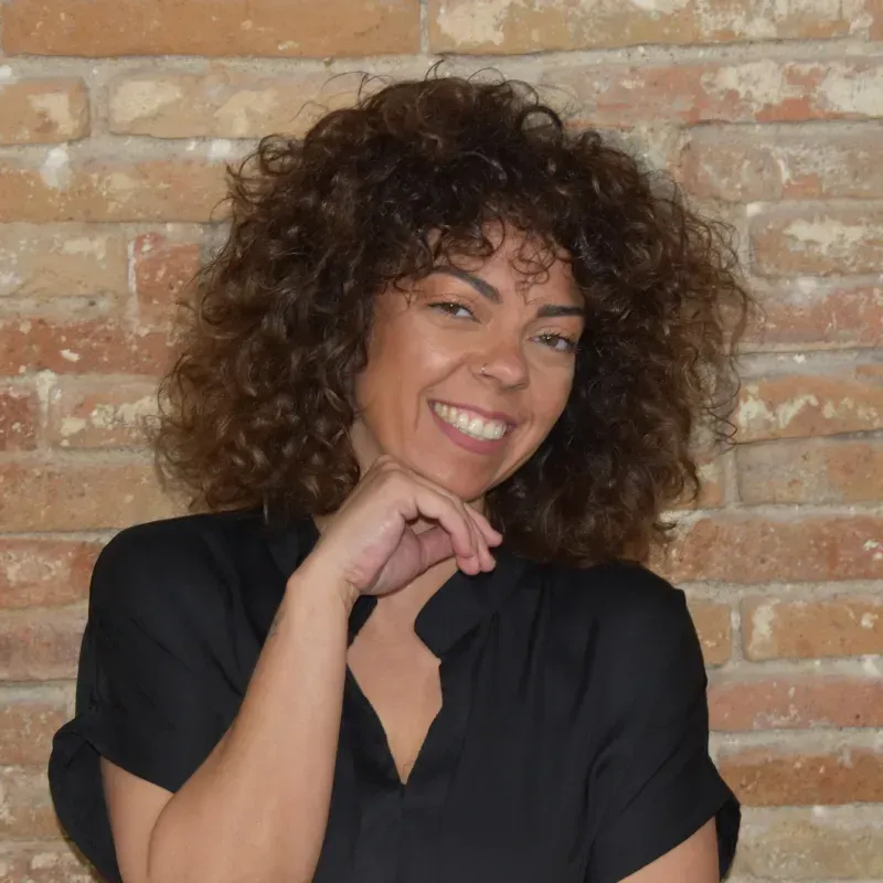 Persona sonriente con cabello rizado y camisa negra, posando frente a una pared de ladrillos.