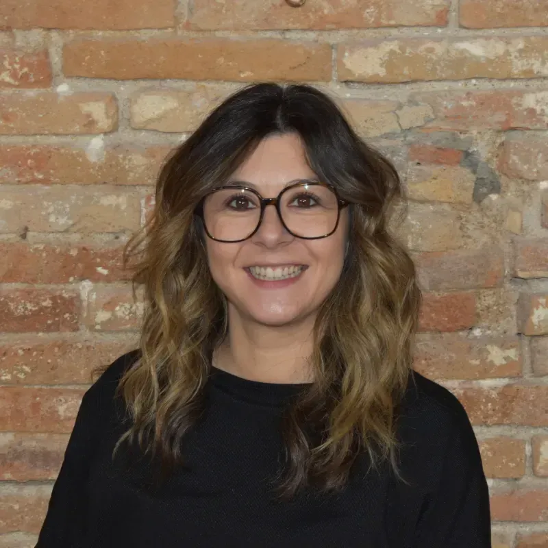Persona sonriente con gafas y cabello ondulado, vestida con una blusa negra, apoyada contra una pared de ladrillos.