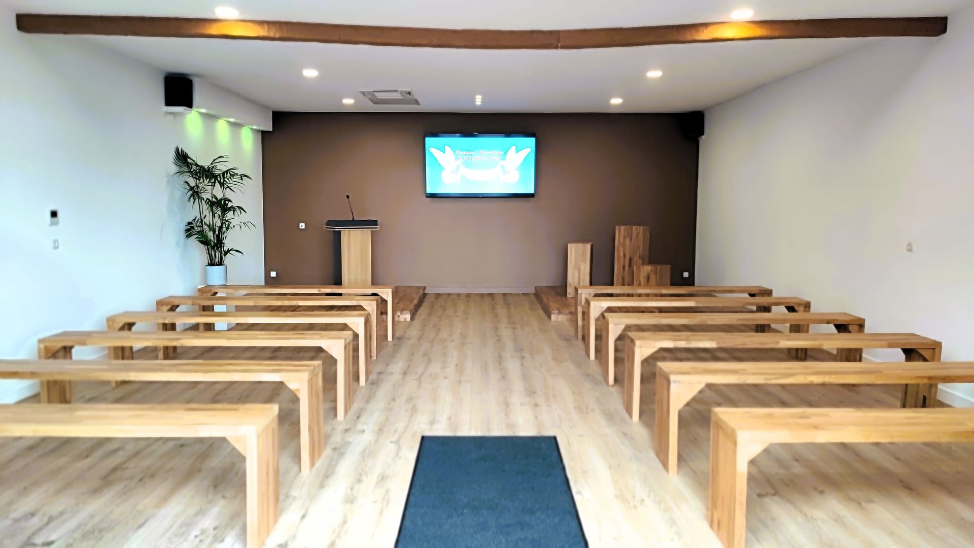 Intérieur d'une salle de cérémonie, avec des bancs, un pupitre et une télé accroché à un mur