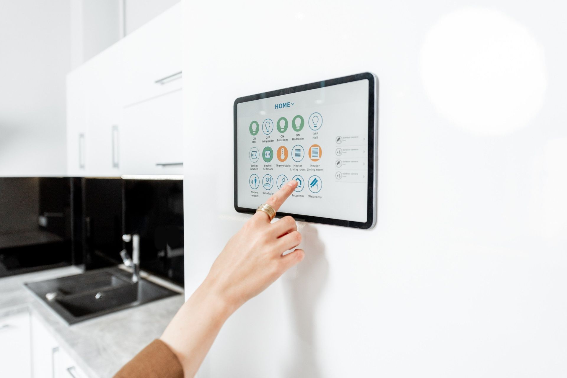La mano de una persona tocando un panel de control de casa inteligente montado en la pared de la cocina, mostrando varios íconos.