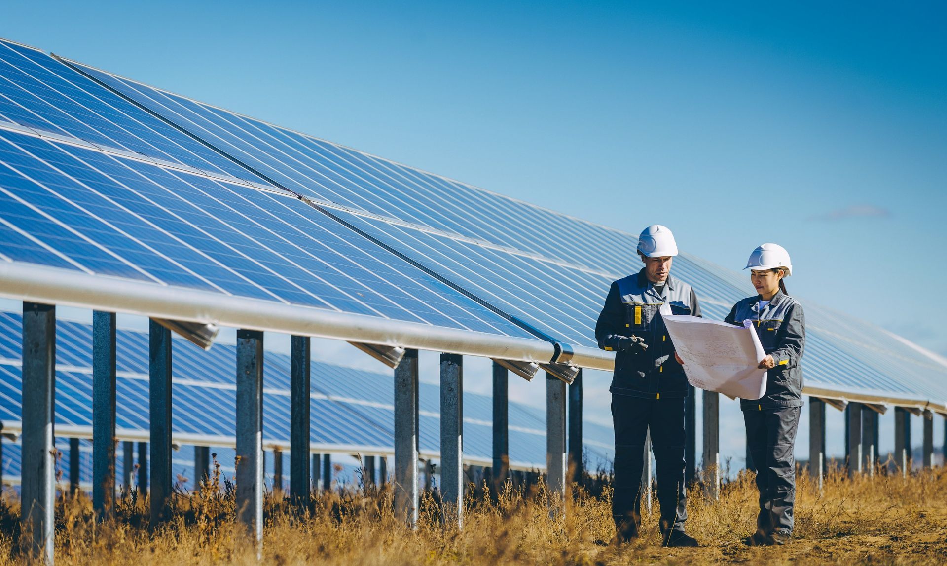 Dos personas con ropa de trabajo y cascos revisando planos frente a un conjunto de paneles solares.