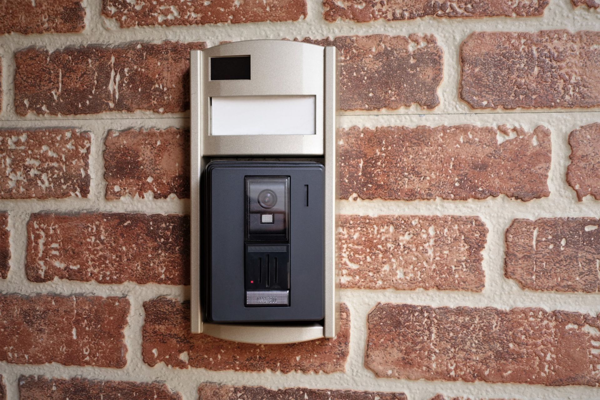 Timbre intercomunicador en una pared de ladrillos. Dispositivo beige y negro con cámara, botón y placa de identificación.