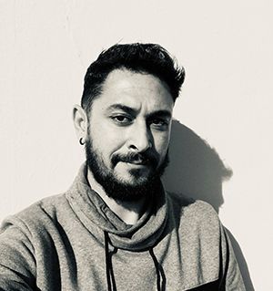Un hombre con barba está parado frente a una pared blanca en una fotografía en blanco y negro.