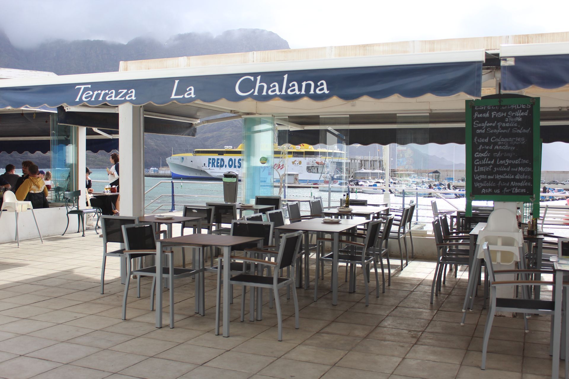 Exterior del restaurante con edificios blancos, toldos, mesas y tablero de menú a lo largo de un camino pavimentado.
