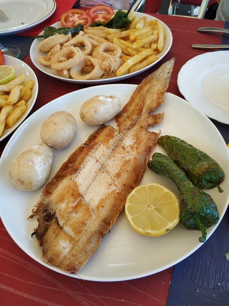 Pescado a la parrilla con patatas, pimientos y limón en un plato blanco, otros platos en la mesa.