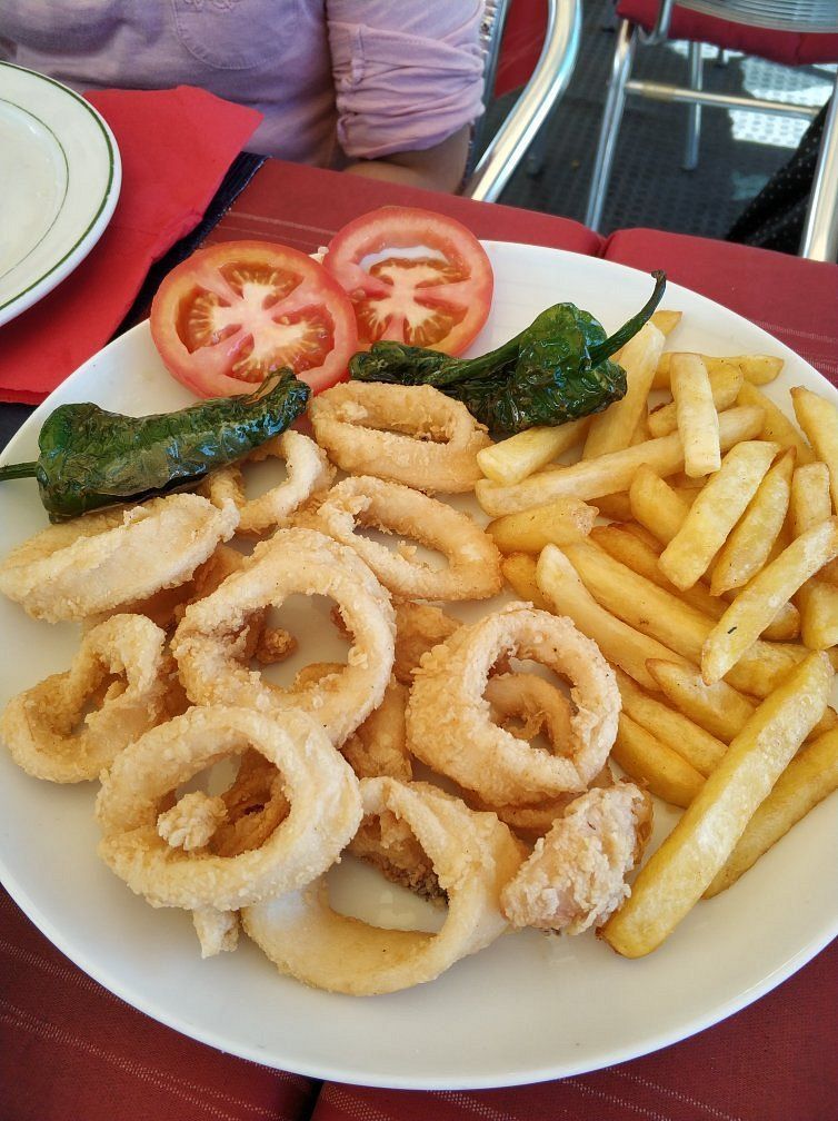Plato de aros de calamares fritos, patatas fritas, rodajas de tomate y pimientos verdes sobre una mesa roja.