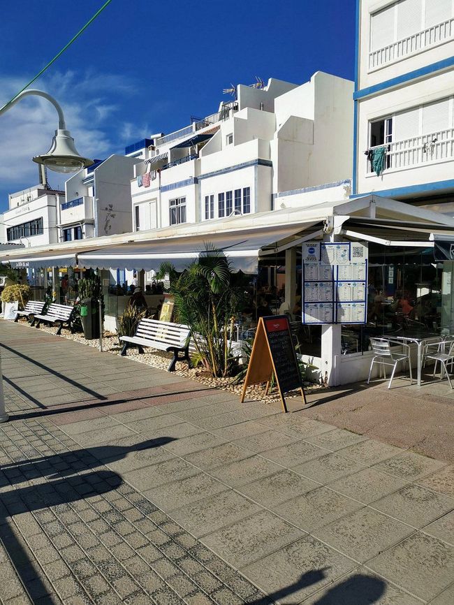 Exterior del restaurante con edificios blancos, toldos, mesas y tablero de menú a lo largo de un camino pavimentado.