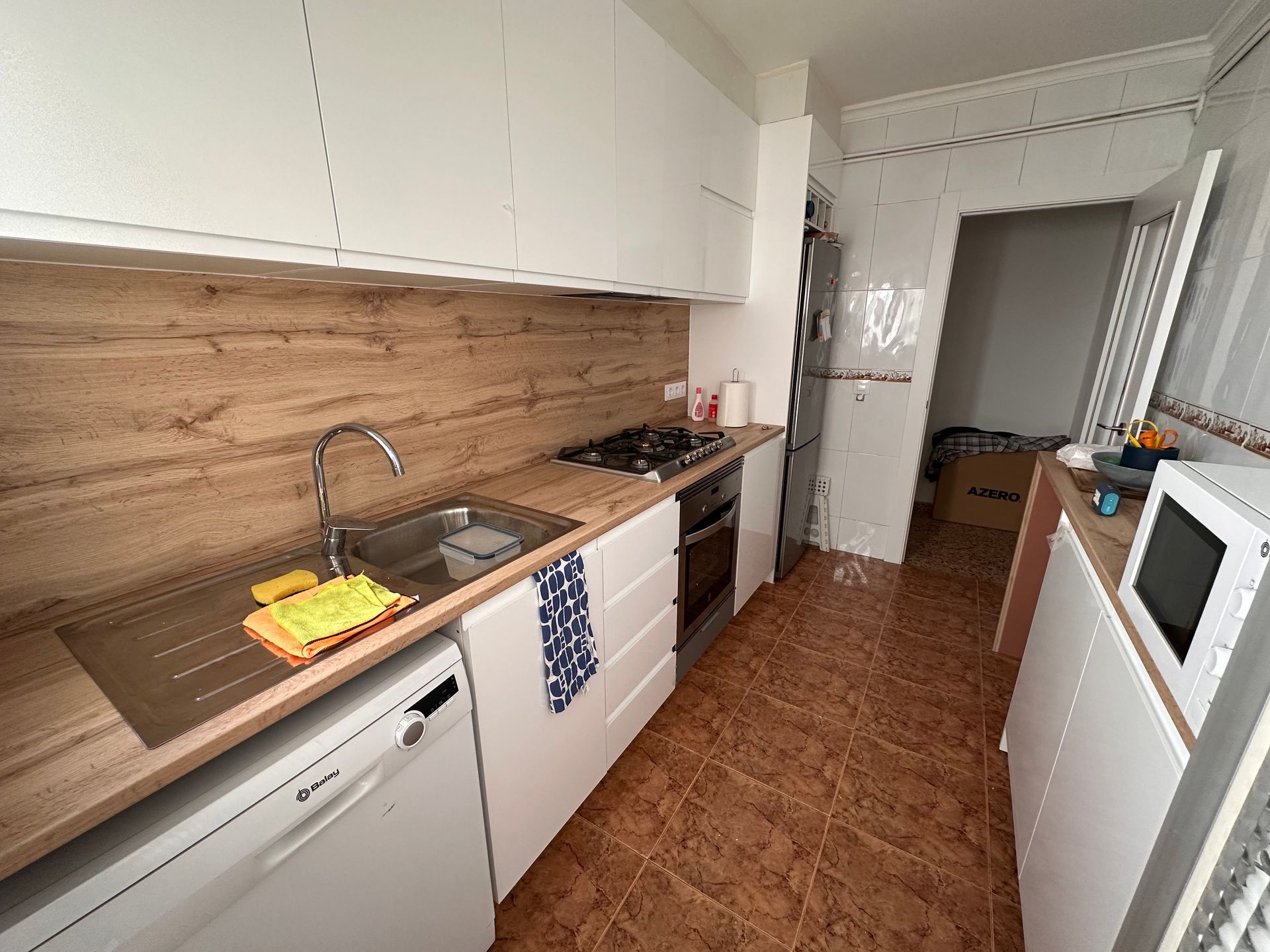 Cocina pequeña con gabinetes blancos, encimeras de madera y electrodomésticos como lavavajillas, horno y microondas.