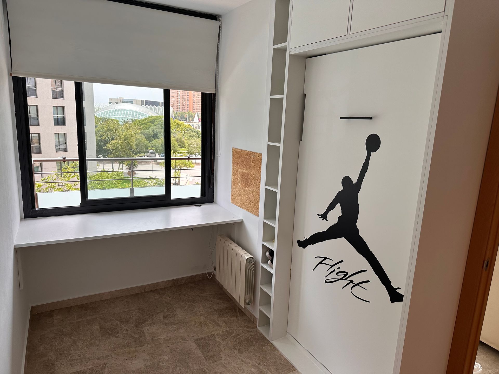 Habitación pequeña con ventana y escritorio, armario de pared con el logo de Michael Jordan y estantería empotrada.