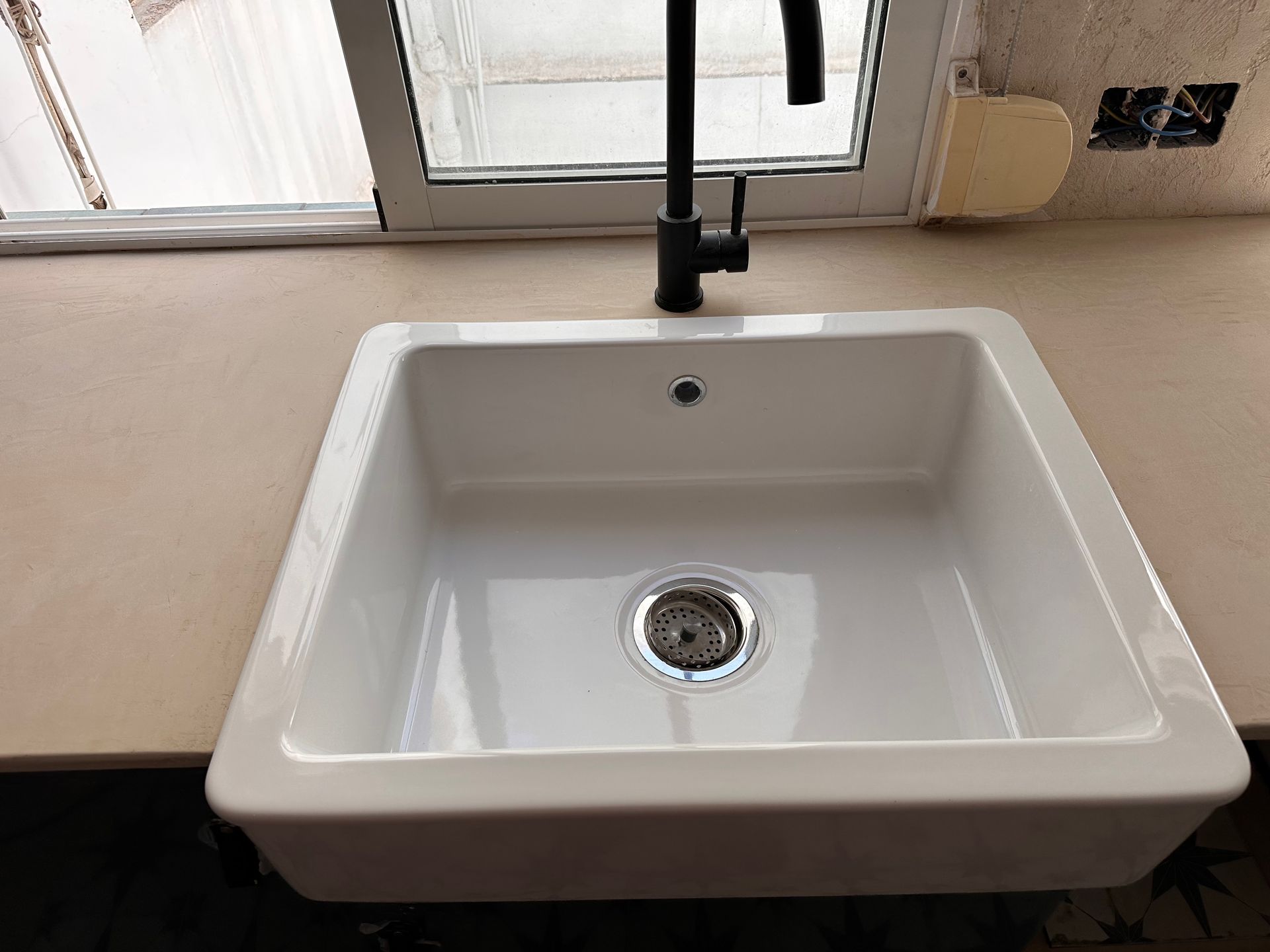 Fregadero de granja blanco con grifo negro, encimera y ventana en el fondo.