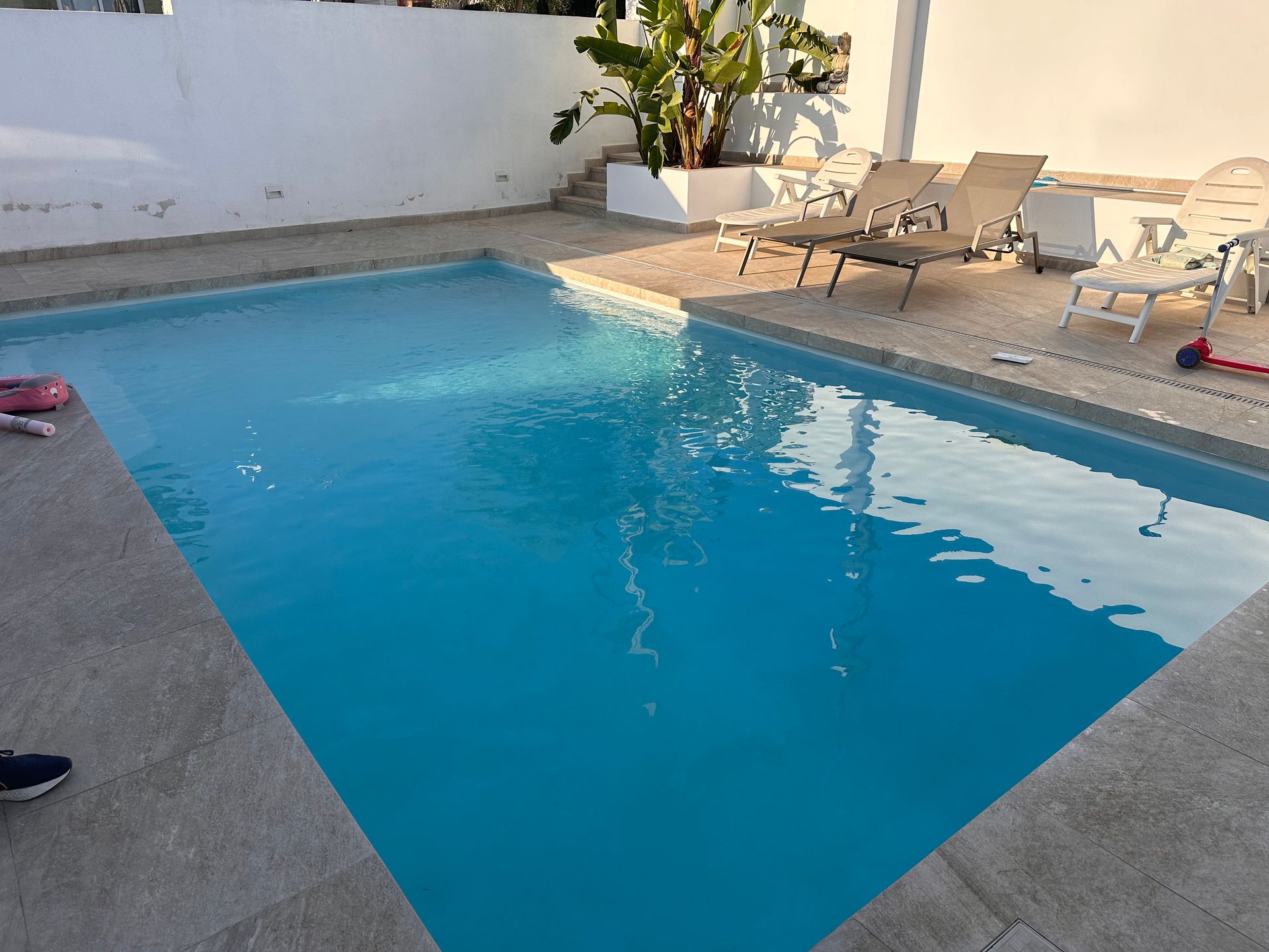 Una piscina rectangular de agua azul rodeada de azulejos beige, tumbonas y una pared blanca.