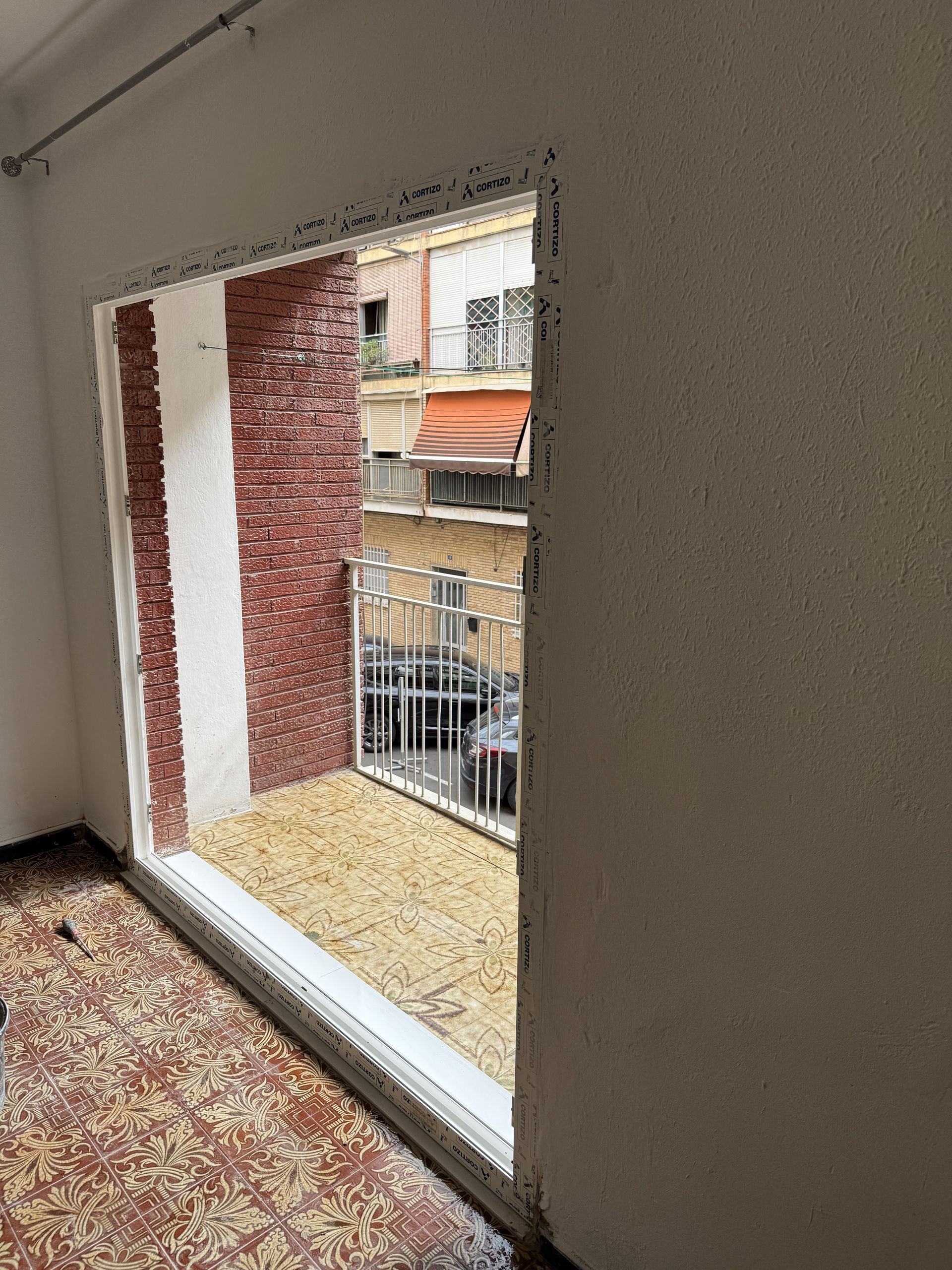 Ventana abierta en un edificio con vista a una calle exterior, una pared de ladrillo y una puerta de seguridad metálica.