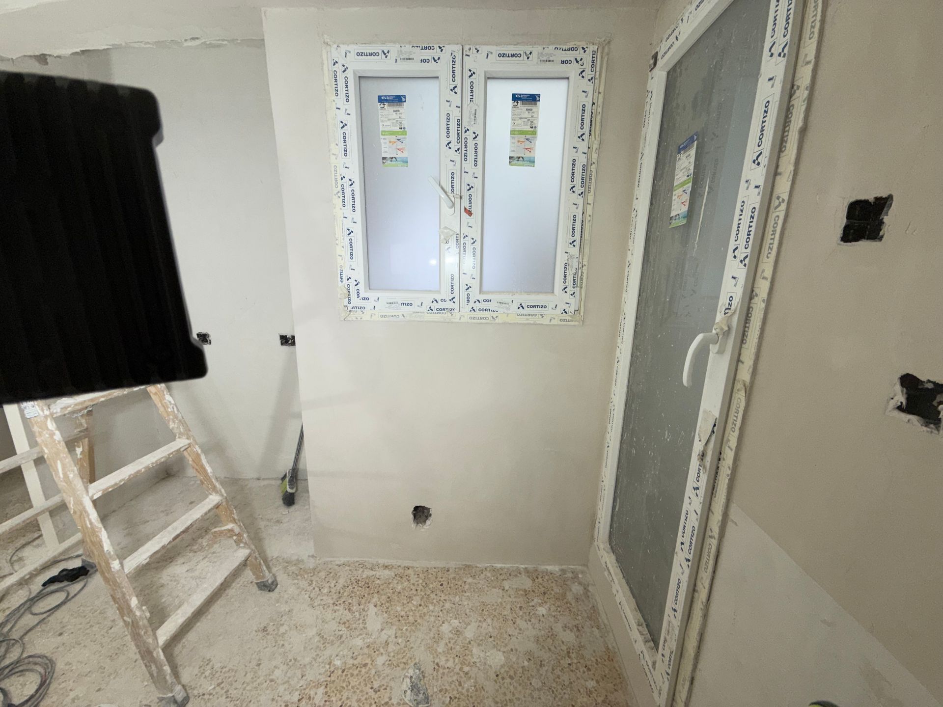 Vista interior: ventana y puerta en una habitación en construcción, con una escalera y un objeto negro a la izquierda.