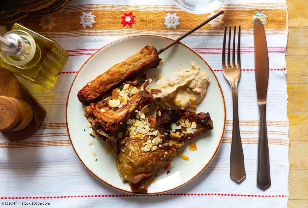 Ein Teller mit gebratenem Hähnchen, dazu ein Spieß und Hummus, auf einem gemusterten Platzdeckchen, daneben Gabel und Messer.