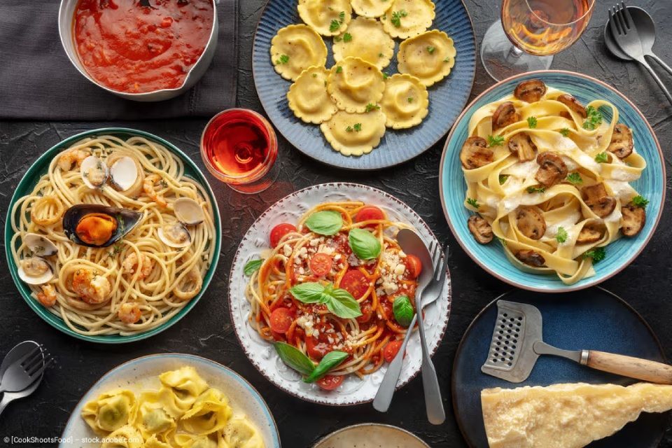 Eine Draufsicht auf einen dunklen Tisch, der mit verschiedenen Schüsseln mit Pasta, einer Schüssel mit roter Soße und einem Stück Parmesankäse gefüllt ist.