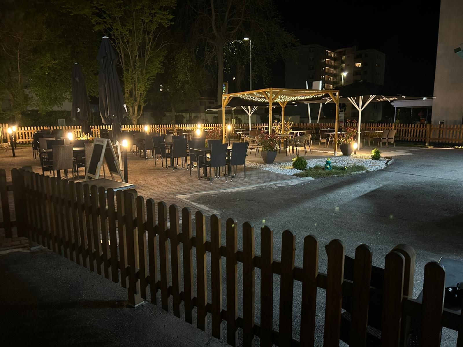 Im Vordergrund steht ein mit Speisen gefüllter Grill, während sich im Hintergrund eine Gruppe von Menschen bei einem sonnigen Essen im Freien unterhält.